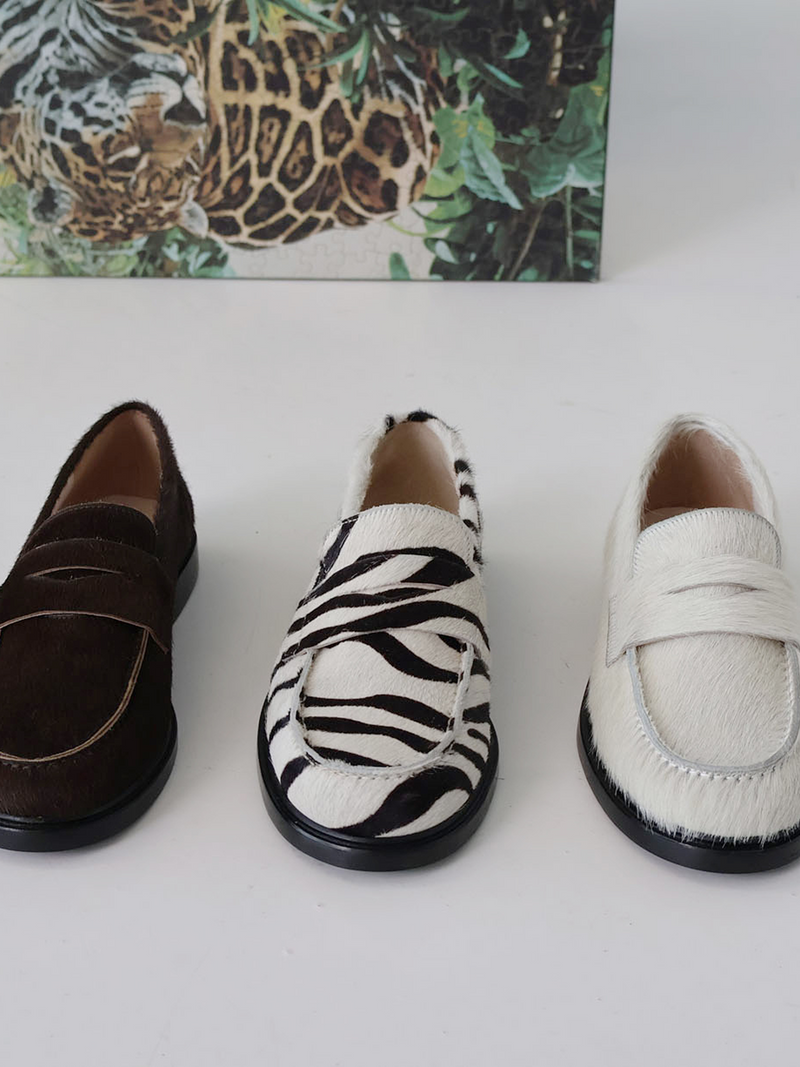 Three Orio loafers in different styles on a white surface with a leopard print box in the background.