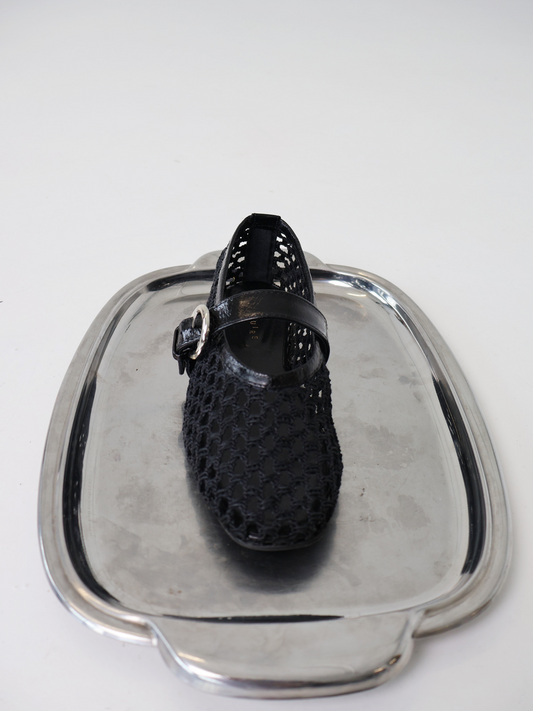 A black woven ballerina flat with an adjustable Mary-Jane strap, displayed on a silver metallic plater