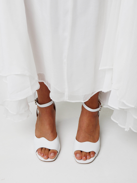 A white heel with an asymmetric wave design and an adjustable ankle strap, made of leather. The are worn with a white wedding dress.
