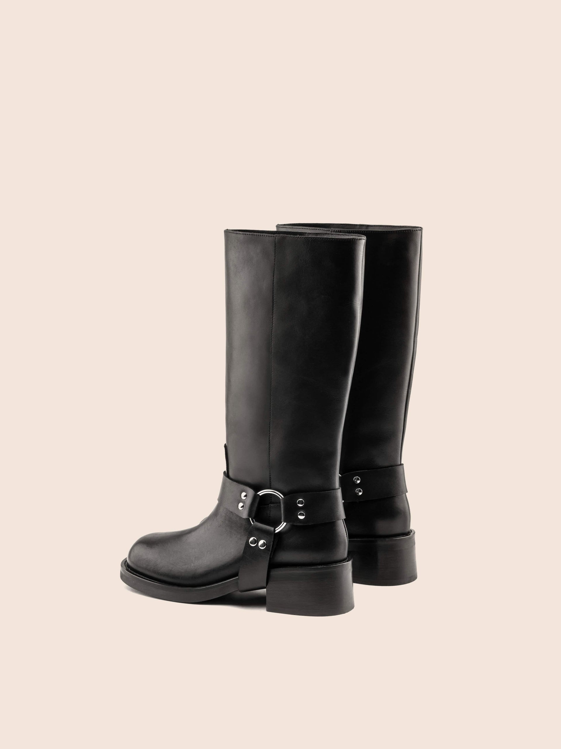 A black leather boot with a harness detail and block heel, on a white background.