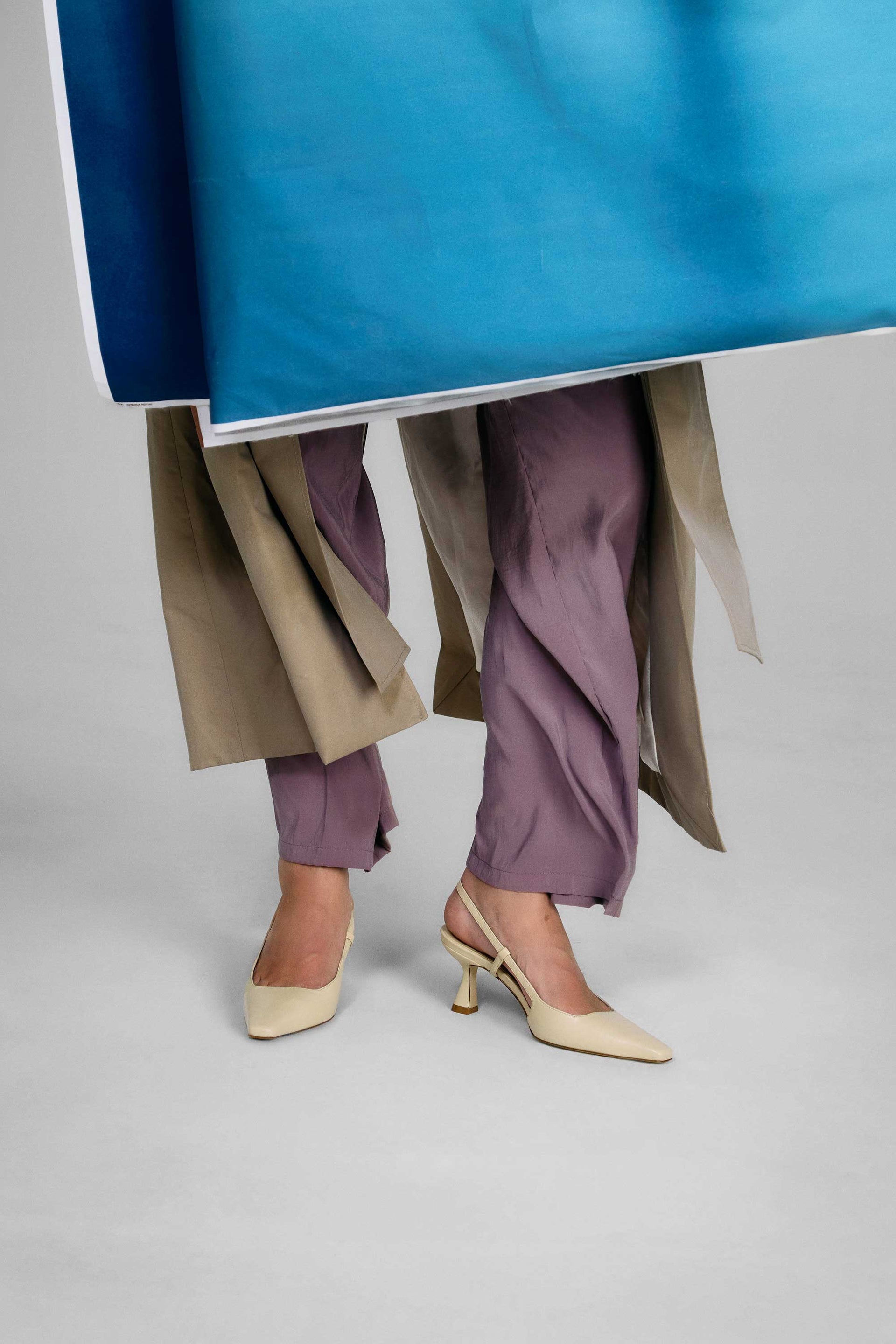 Person wearing a blue top, beige coat, purple pants, and beige shoes on a gray background