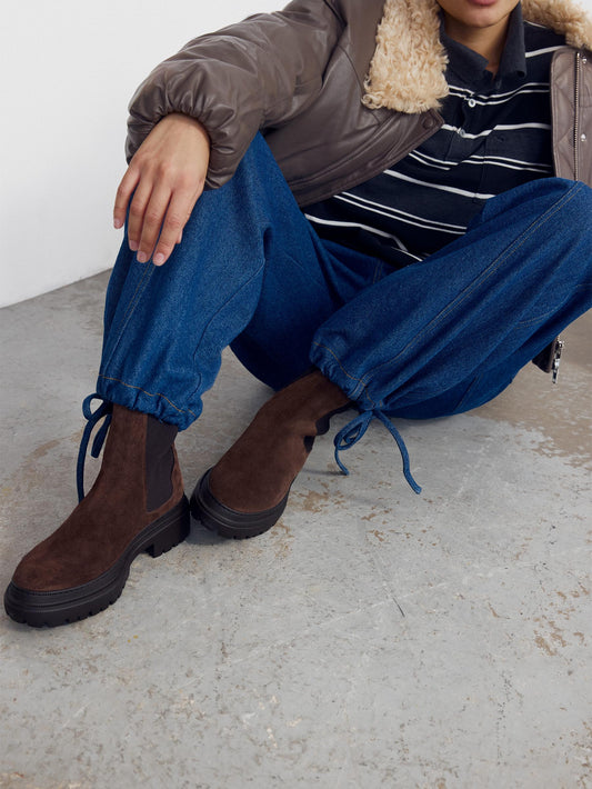 Woman wearing brown suede chelsea boots, sitting on ground and wearing some balloon pant jeans with a striped shirt and pleather brown jacket.