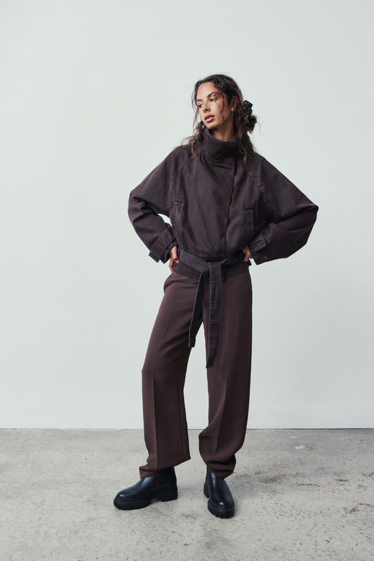 Person wearing a dark outfit with a high collar and wide-leg pants against a plain background