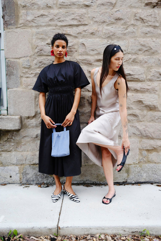 Two women standing against a stone wall, one in a black dress and the other in a beige dress.
