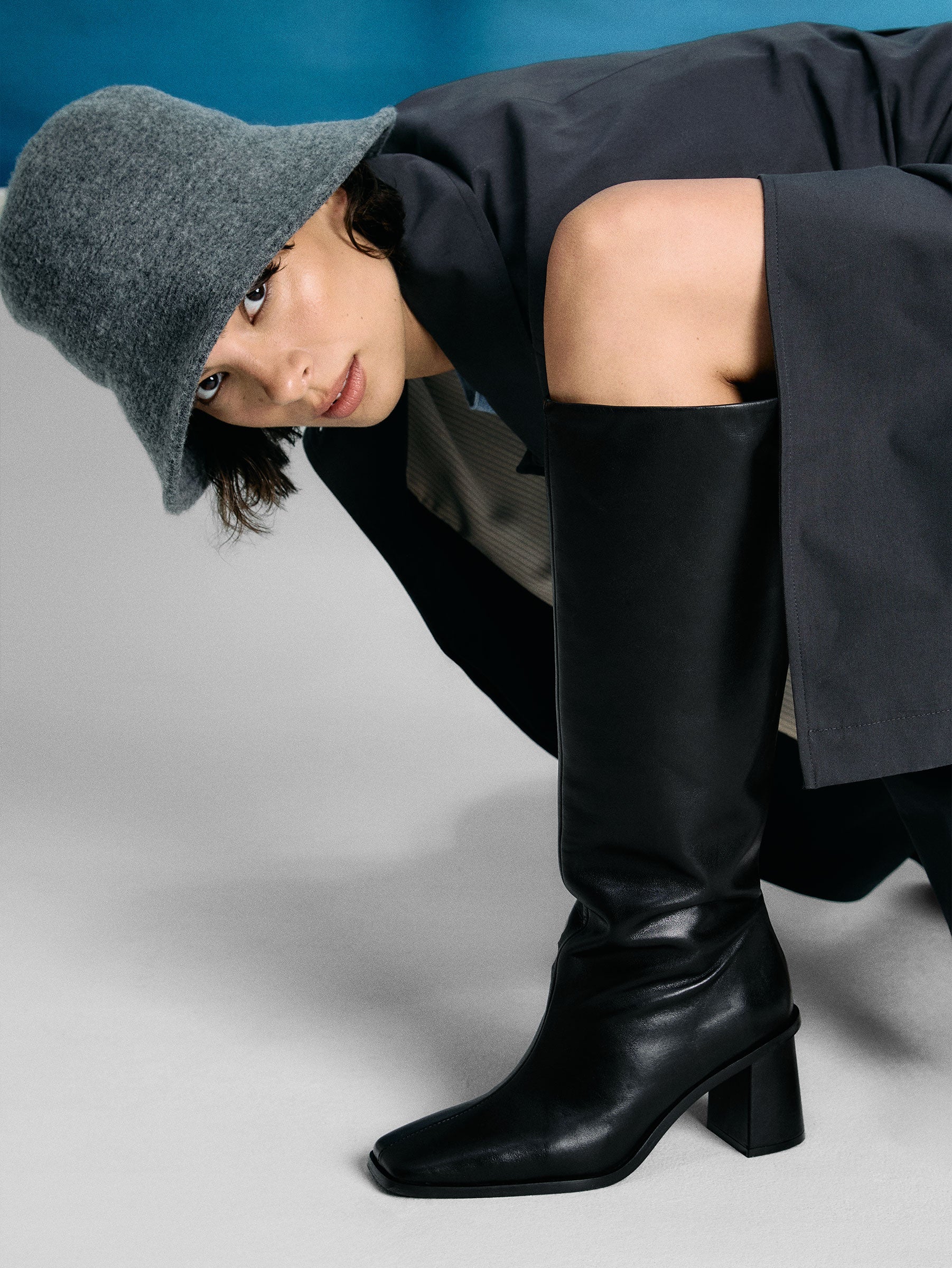Person wearing a gray beanie, black coat, and black knee-high boots on a gray background