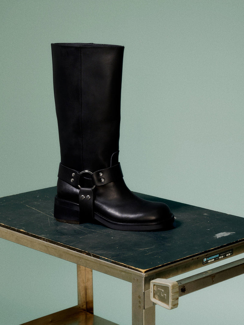 A black leather boot with a harness detail and block heel, on a green background.