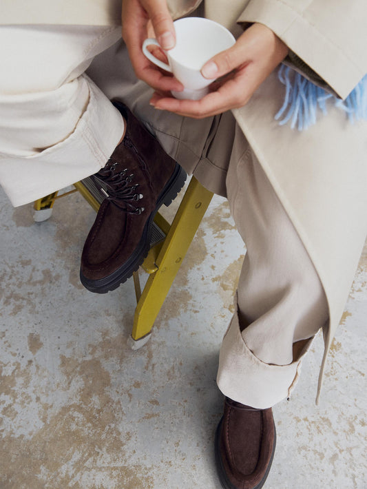 A brown suede chunky combat boot with lace-up closure and metal hardware worn with nude pants