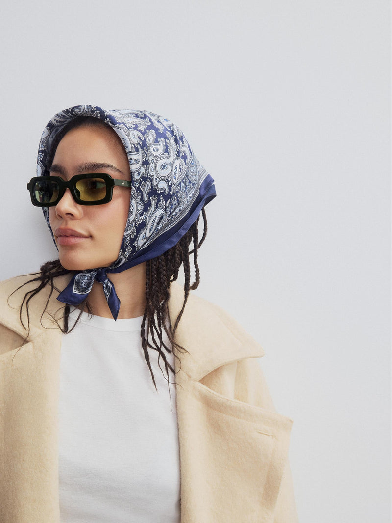 Woman wearing rectangle shaped sunglasses in a dark green colour with a lighter green lens. Wearing a blue paisly scarf on her head. 
