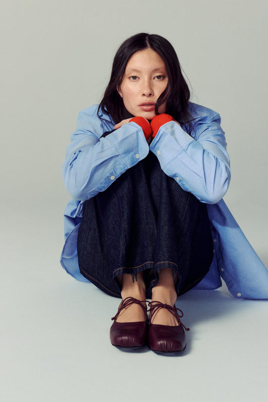 woman sitting on white floor against same coloured background, wearing dark wash jeans, with a blue shirt and plum coloured ballet flats with lace bow detailing