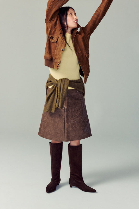 girl standing in white studio wearing brown suede heeled boots, brown skirt, yellow top with green sweater at her waist, smiling and playing around with jacket