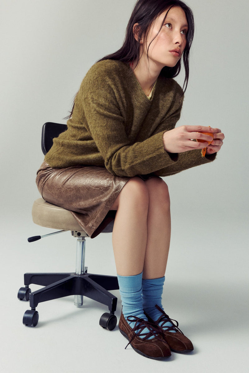 woman sitting on rolling chair wearing brown skirt, green sweater & blue socks
