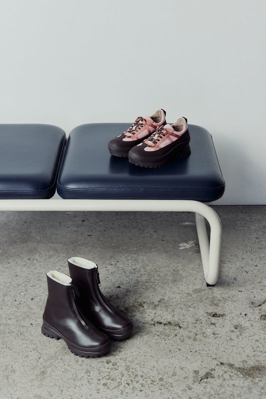Pair of dk brown winter boots on a concrete floor with a bench in the background. The pink sneaker are on the bench.