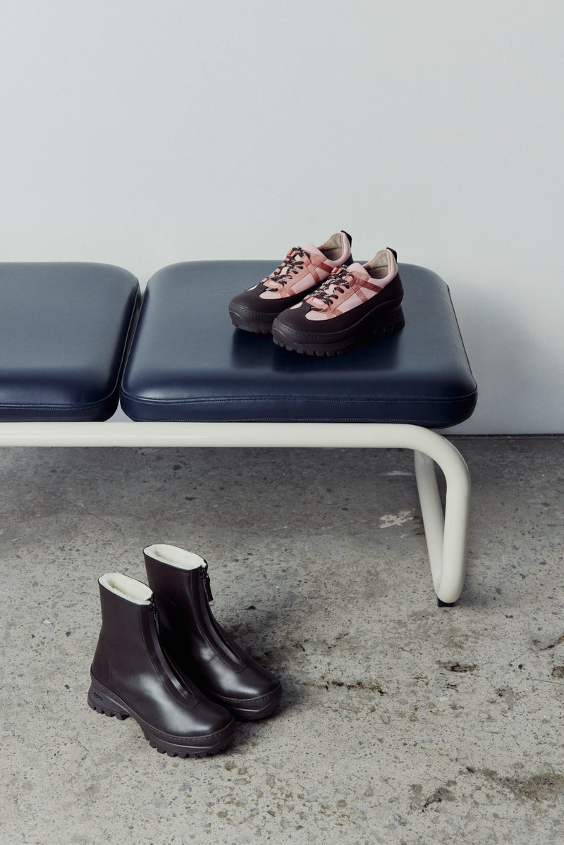 Pair of dk brown winter boots on a concrete floor with a bench in the background. The pink sneaker are on the bench.