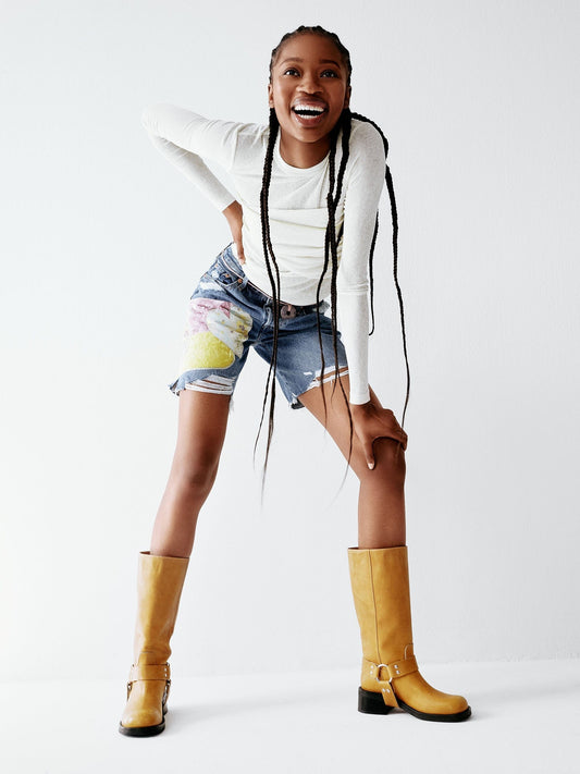 A yellow tan leather boot with a harness detail and low heel worn by a model wearing short.