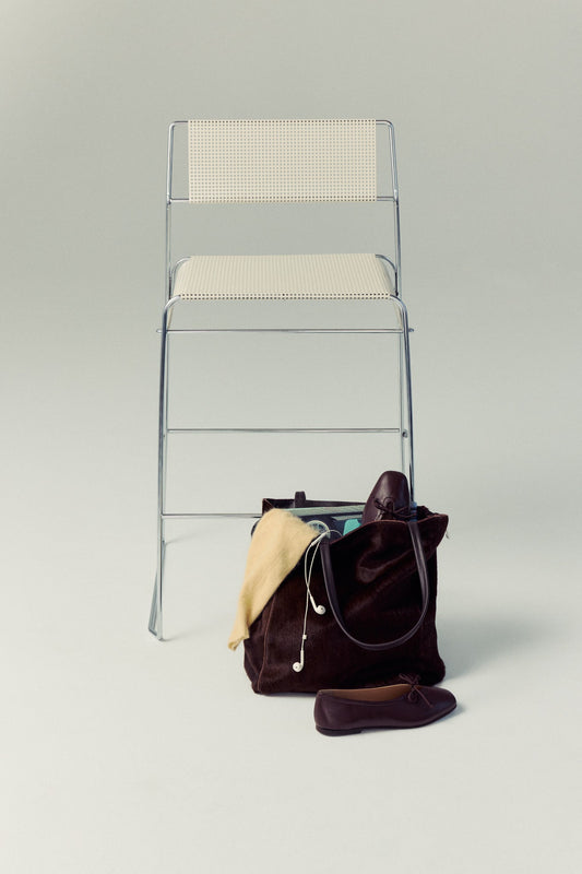 Clear chair with a beige cushion next to a brown Carpi handbag on a light gray background