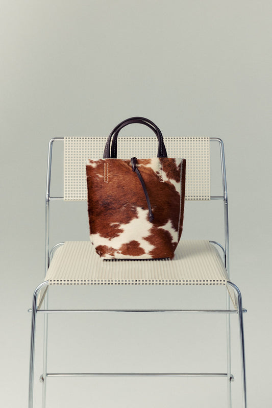 Brown and white cowhide Carpi leather bag on a clear chair with a gray background