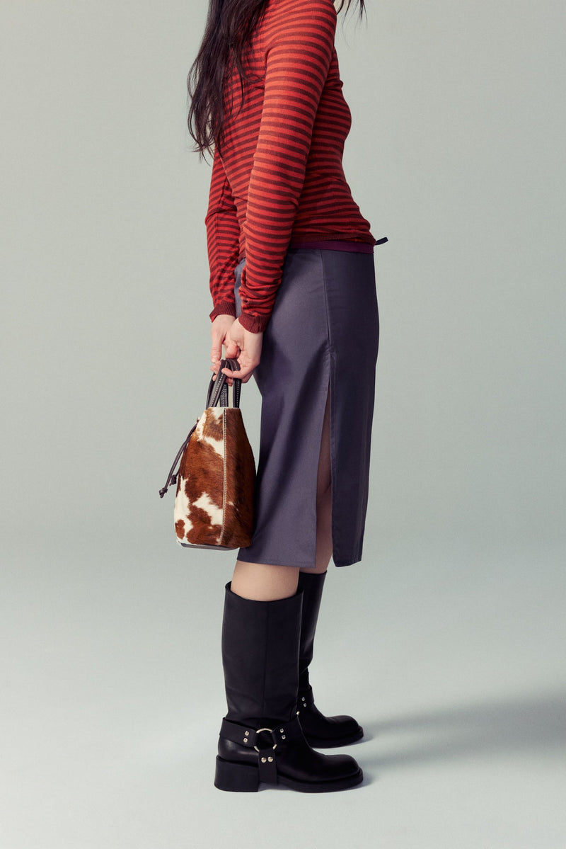 Person wearing a red striped sweater, purple skirt, and black Lucca boots holding a cowhide handbag on a gray background