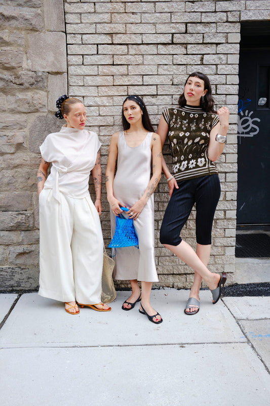 Three women standing against a stone wall, wearing stylish outfits.