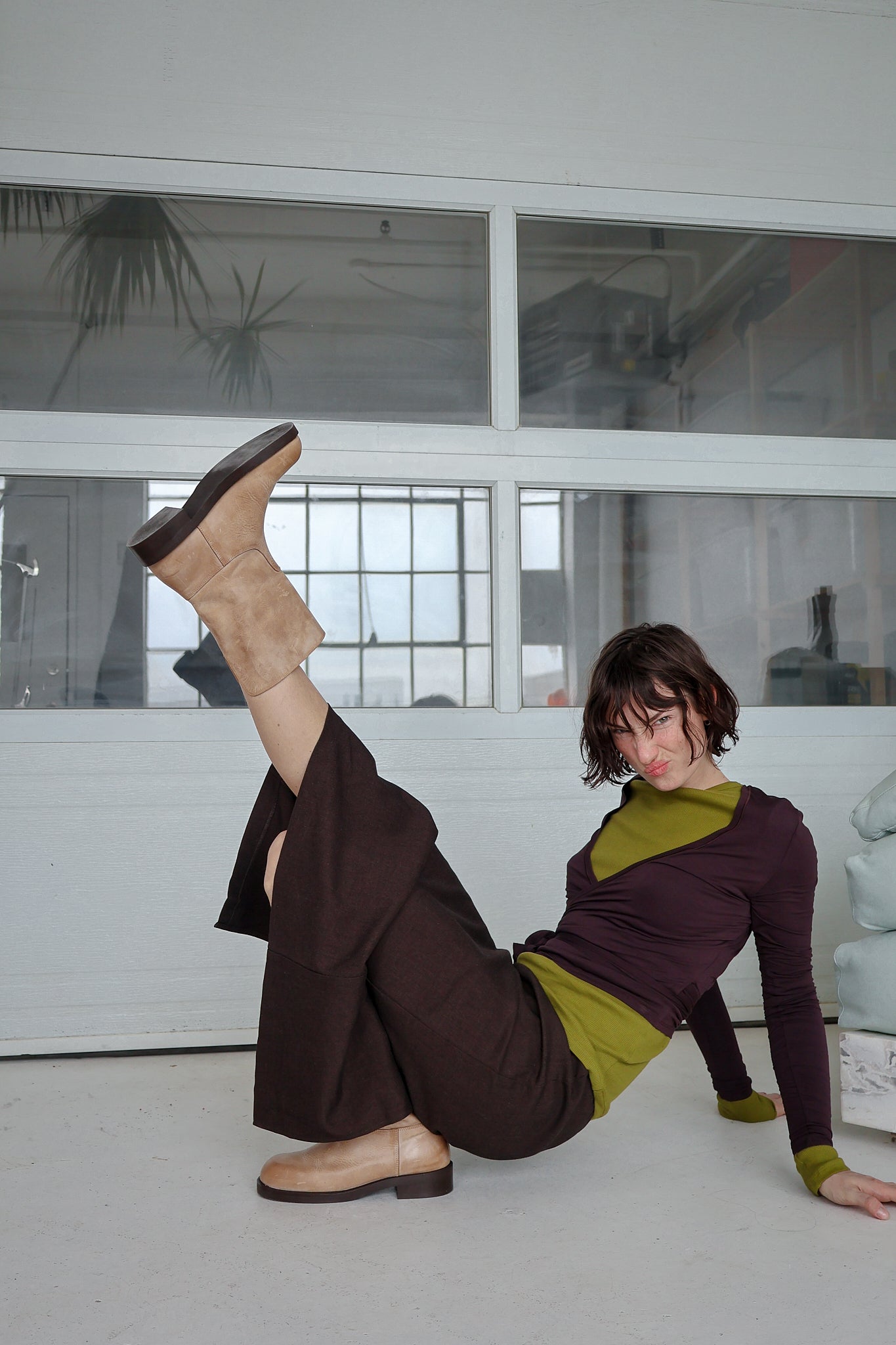 Person sitting on the floor with one leg raised wearing tan boots in a room with large windows.