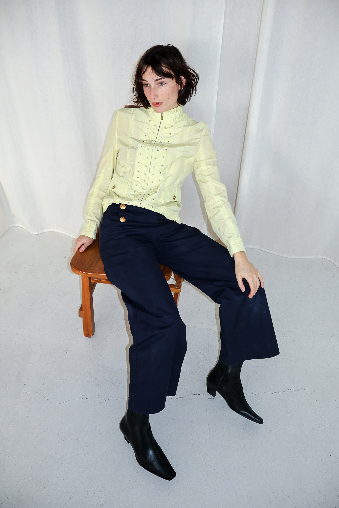 Woman sitting on a stool wearing a light green sweater and dark blue pants against a white background