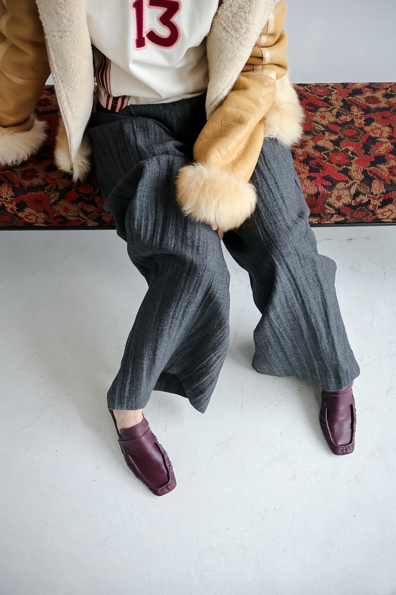 Person wearing a coat with fur trim, white shirt with red number 13, gray ribbed pants, and bordeaux shoes on a white floor.