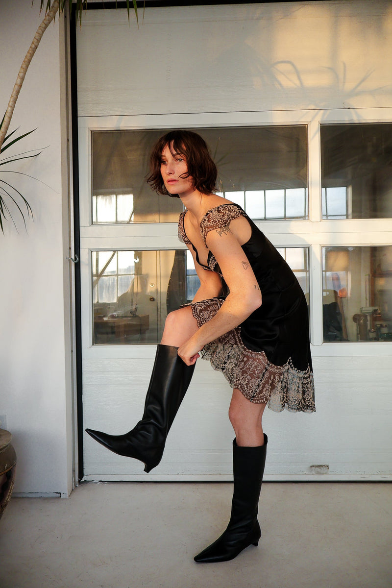 Woman in black lace dress and knee-high boots posing indoors.