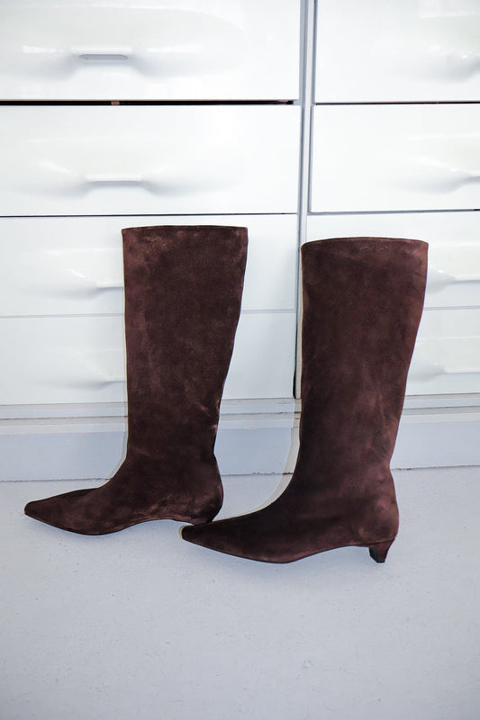 Knee-high brown suede boots with a low heel and pointed toes in front of white dresser