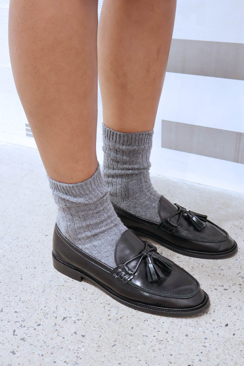 Black loafers with tassels worn with gray socks on a light background