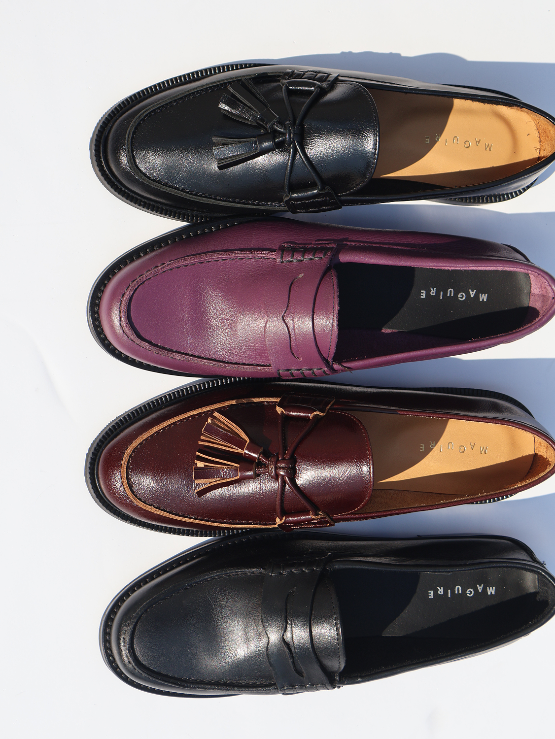 Four pairs of loafers in black, purple, brown, and another black color on a white background.