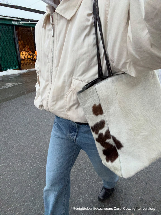 woman wears blue jeans, cream jacket and a cow printed tote bag mostly white, walking through a dimly lit city