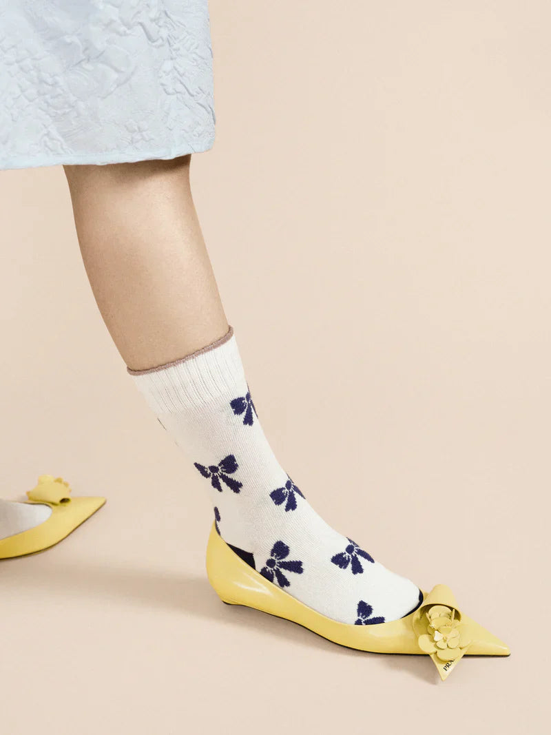 A pair of ivory crew socks with blue bows and yellow shoes, worn on a mannequin with a lacey blue dress .