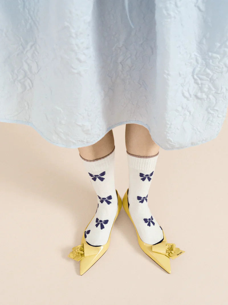 A pair of ivory crew socks with blue bows and yellow shoes, worn on a mannequin with a lacey blue dress .
