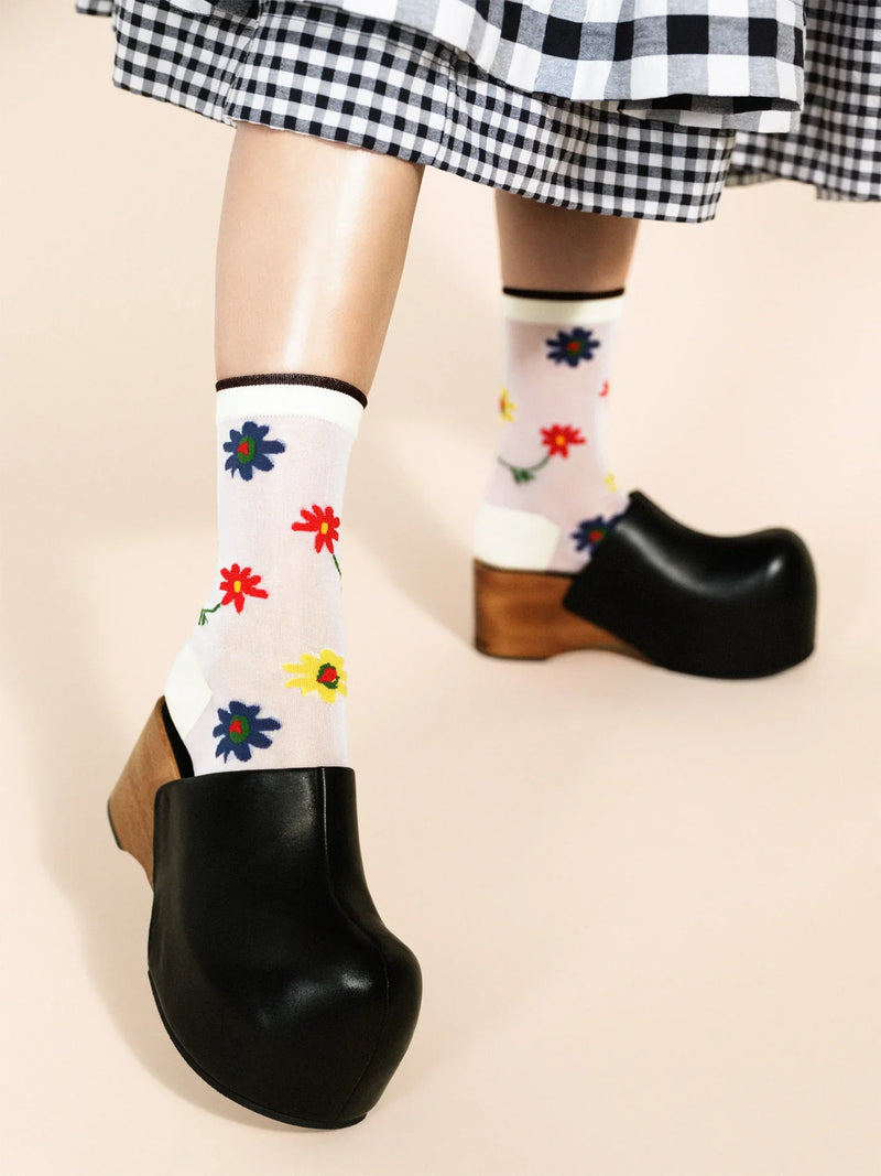 A pair of white socks with a floral pattern worn on feet wearing black shoes, with a checkered skirt visible above.